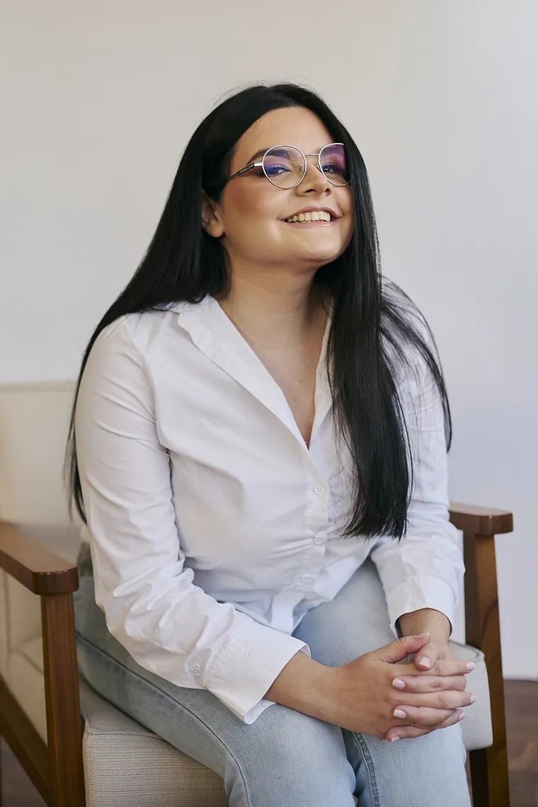 Jennifer da Luz Silva, psicóloga clínica, sorrindo, sentada em uma poltrona, de camisa branca