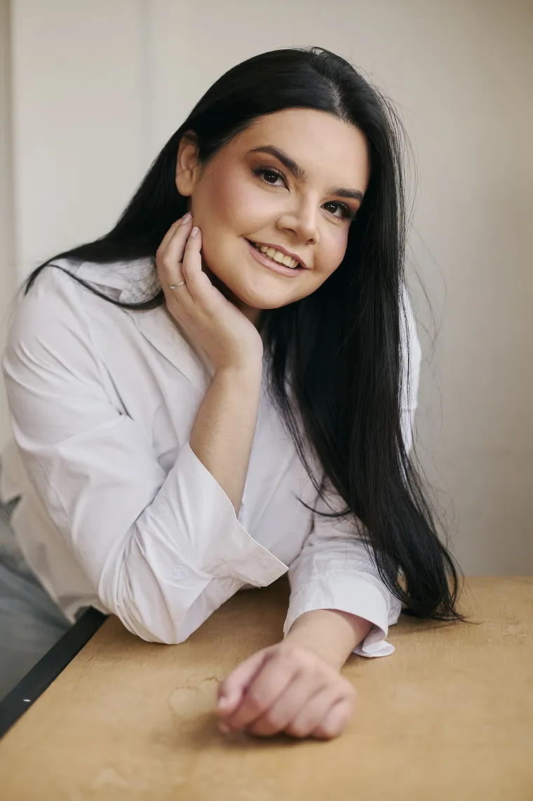 Jennifer da Luz Silva sorrindo, apoiada em uma mesa de madeira, com a mão no rosto
